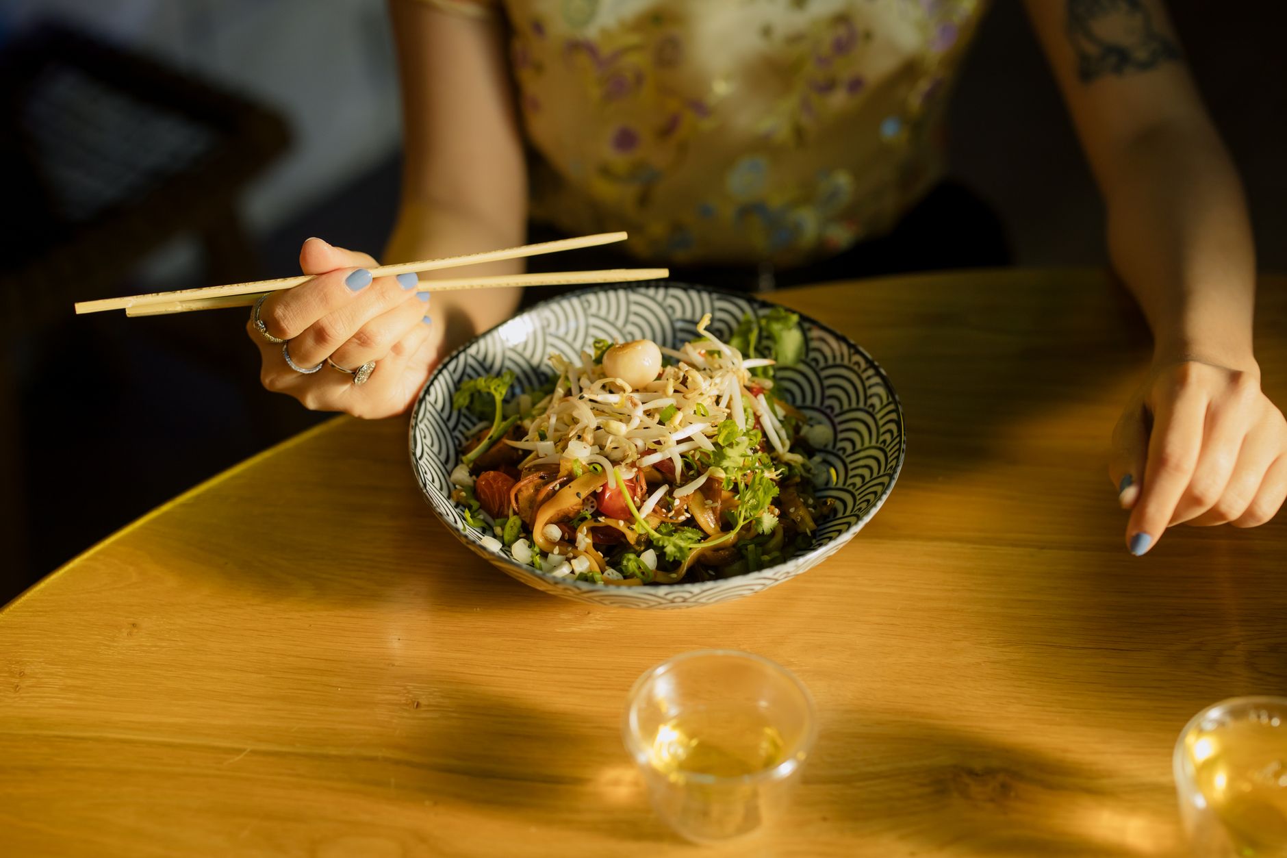 Personne tenant des baguettes au-dessus d’un bol de nouilles aux germes de soja et aux herbes, sur une table en bois, avec deux petits verres au premier plan.